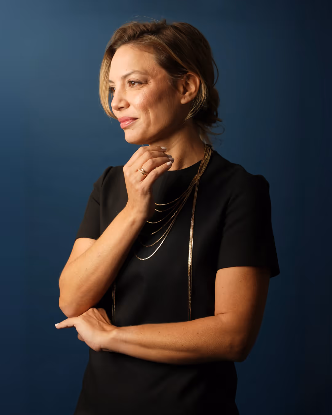 Pamela in black dress with gold necklace and ring looking thoughtfully to the side against a blue background.