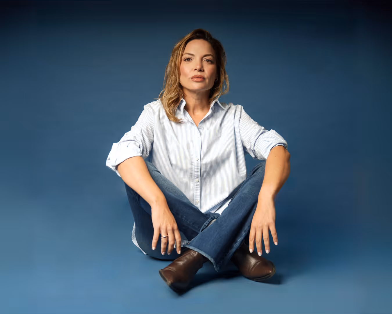 Woman with light brown hair wearing jeans and a white shirt sitting cross-legged on blue background.