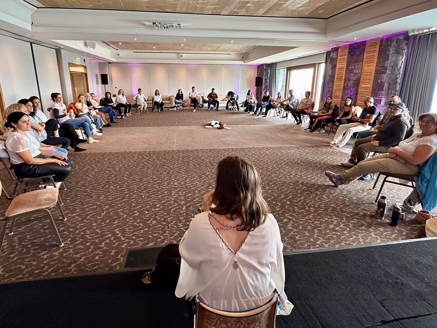 Group of people seated in a large circle in a carpeted conference room with purple lighting.