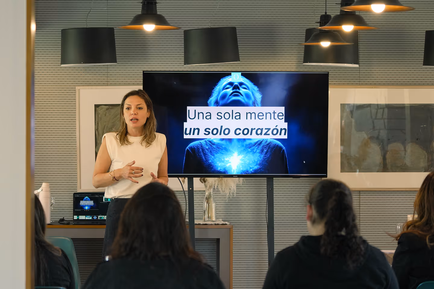 Woman giving presentation to seated audience with screen showing 'Una sola mente, un solo corazón'.