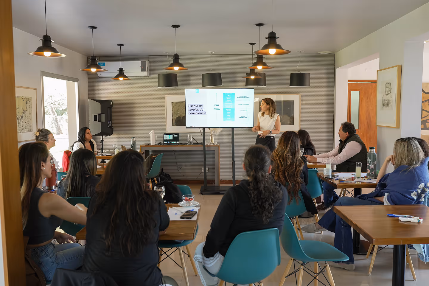 Woman giving a presentation to a group seated at tables in a well-lit room with hanging lights.