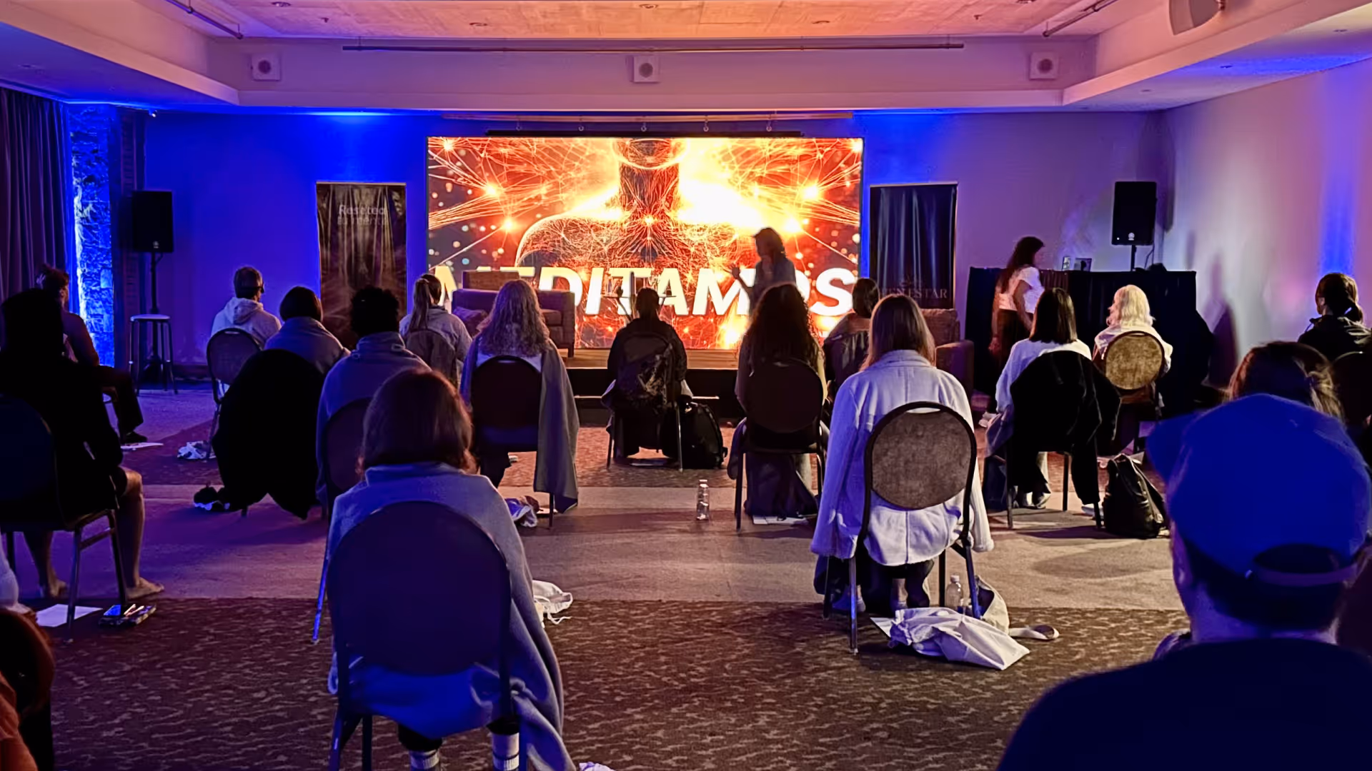 People seated on chairs in a dim room facing a large digital screen with the word MEDITAMOS.