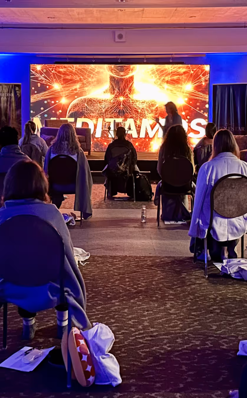 People seated in chairs facing a stage with a glowing digital human figure and the word MEDITAMOS.