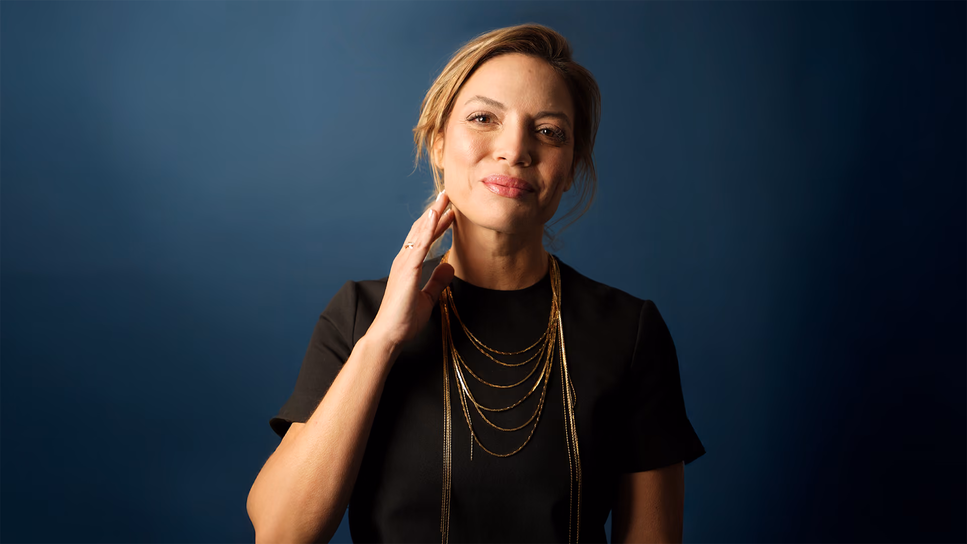 Smiling woman with gold necklaces and black dress against a dark blue background.