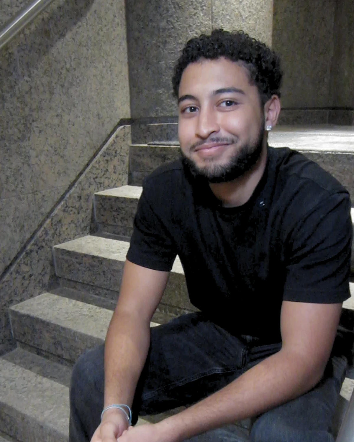 Smiling young man with curly hair and beard, wearing a black shirt and jeans, sitting on stone stairs.