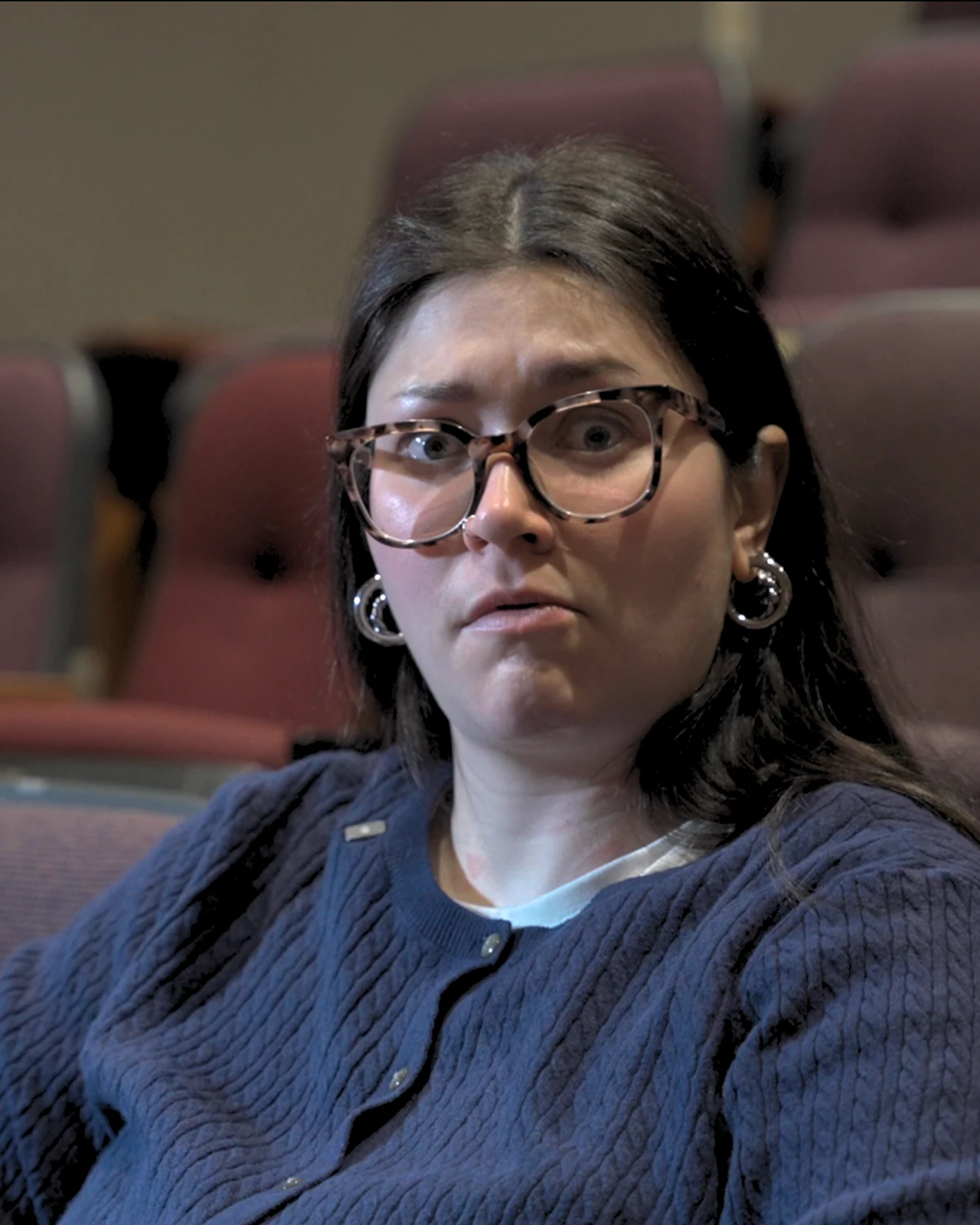 Woman with dark hair, large tortoiseshell glasses, and hoop earrings, wearing a blue cardigan, sitting in a theater with surprised expression.