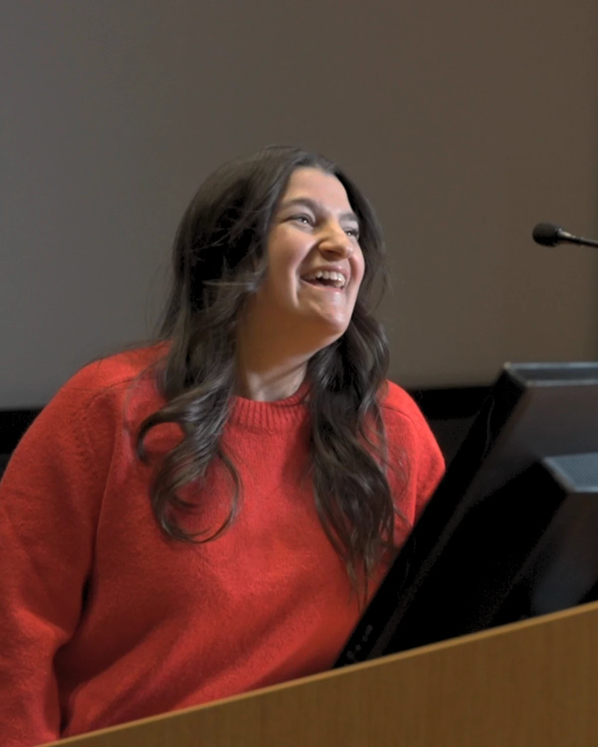 Smiling woman with long dark hair in a red sweater speaking or presenting behind a podium with a microphone.
