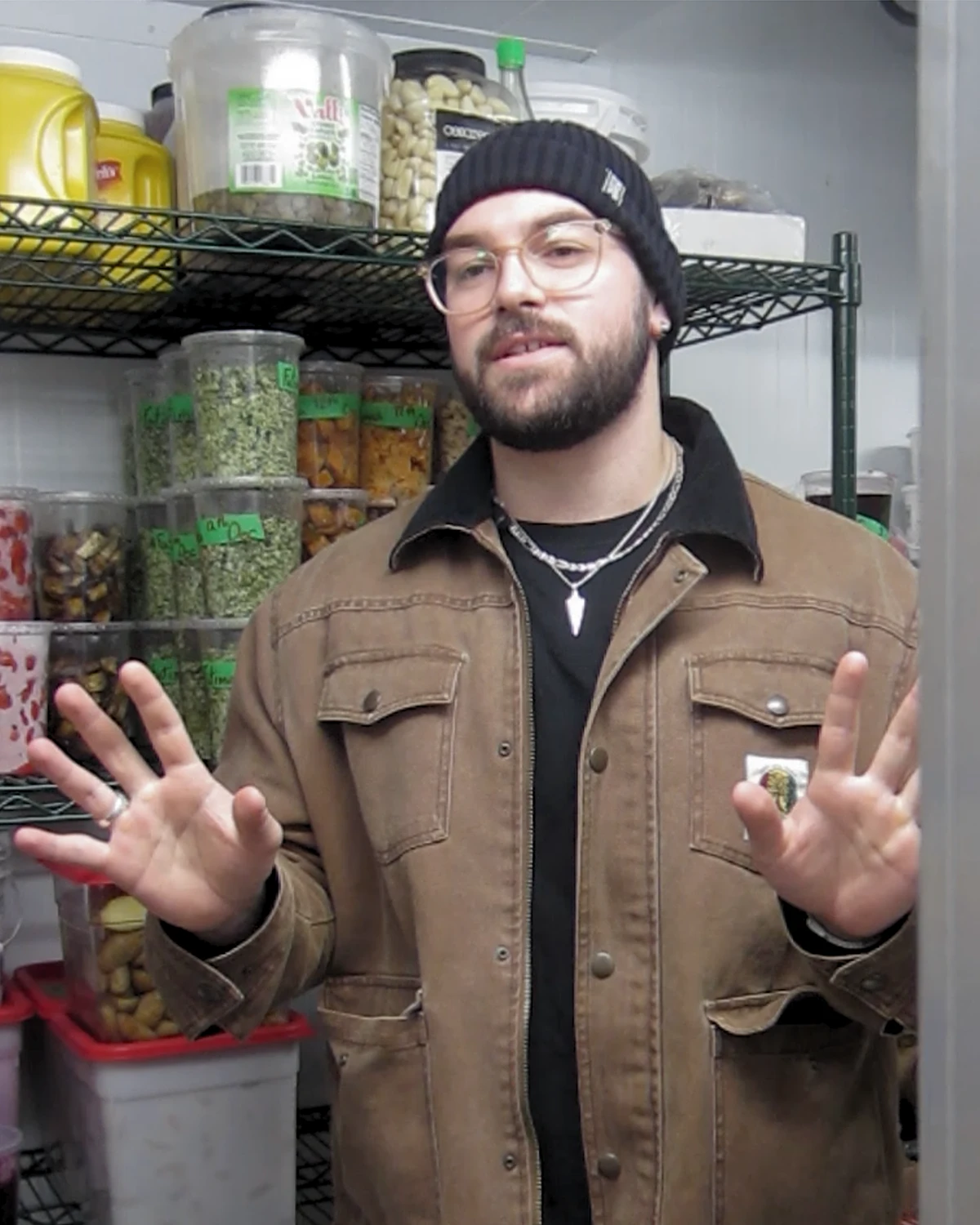 Man wearing glasses, black beanie, brown jacket, and necklaces standing with hands raised in front of shelves filled with containers of various food items.