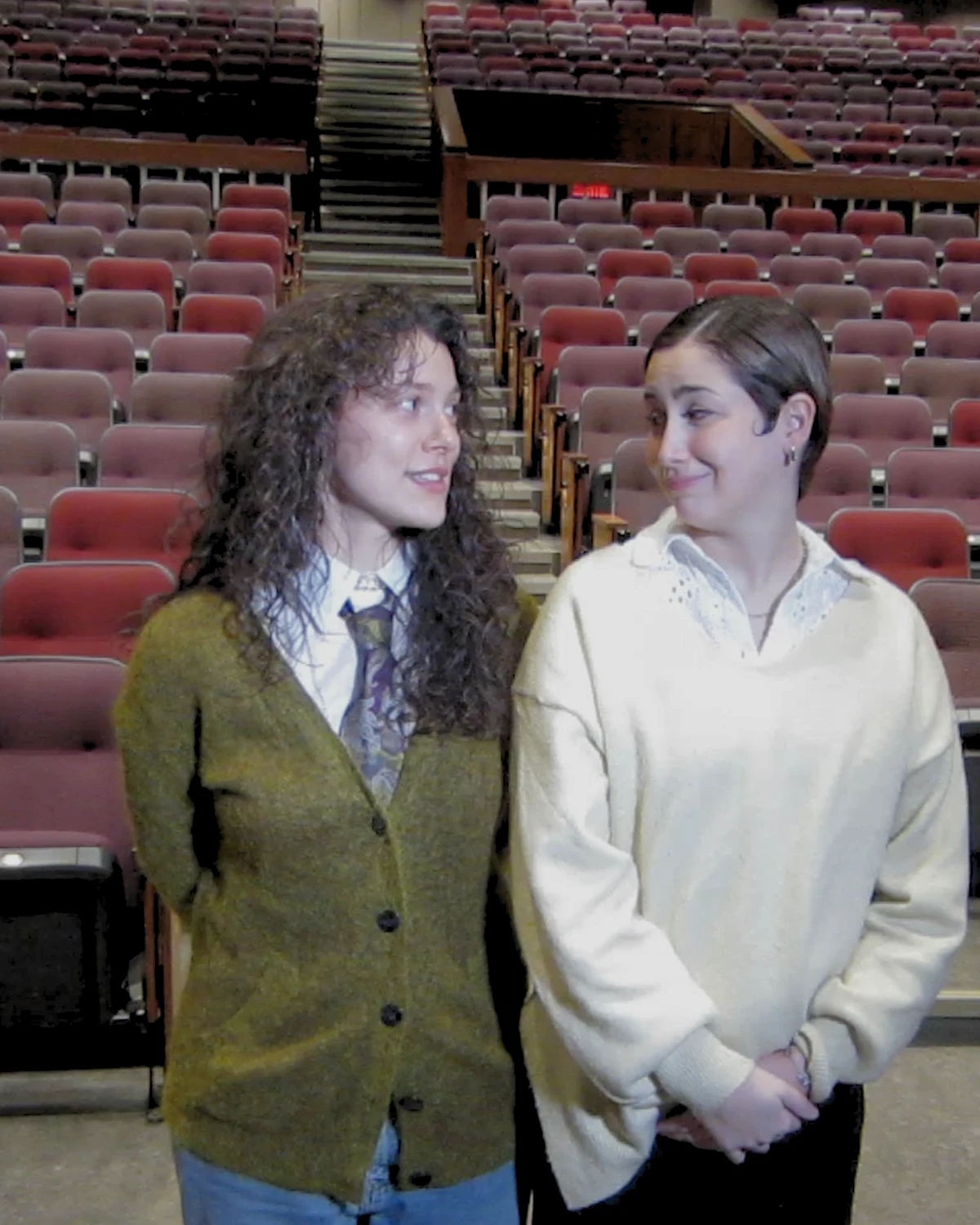 Two young women standing side by side in a large auditorium with empty red seats, looking at each other and smiling.
