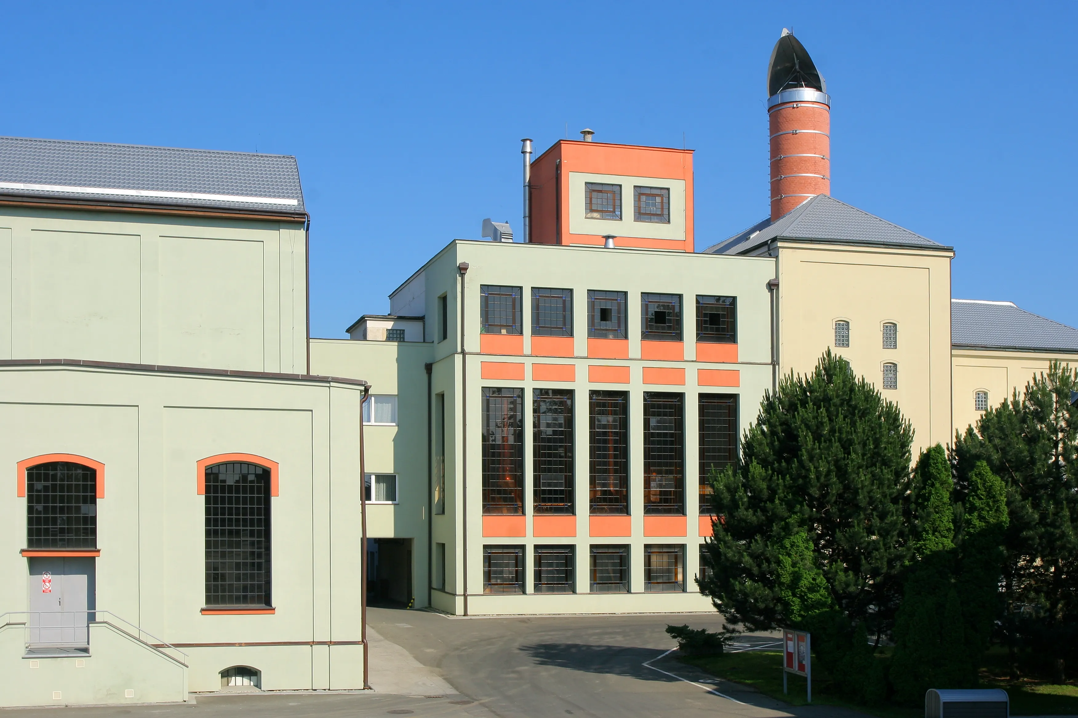 Moderní průmyslová budova s oranžovými a světle zelenými detaily, vysokým komínem a několika okny za jasného modrého nebe.