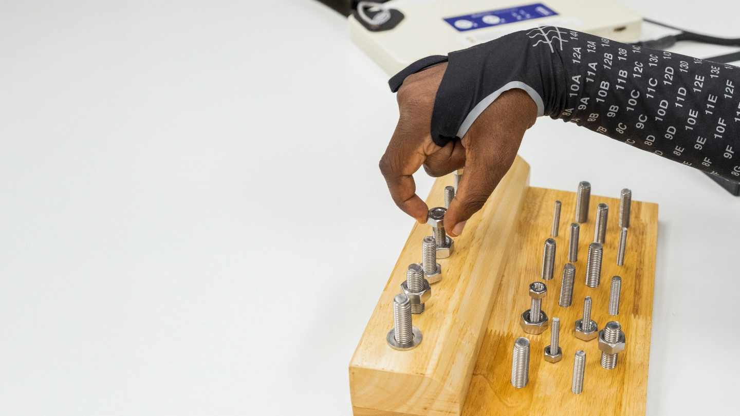 Man wearing a black sensor-covered sleeve exercises fingers with metal pegs on a wooden board.