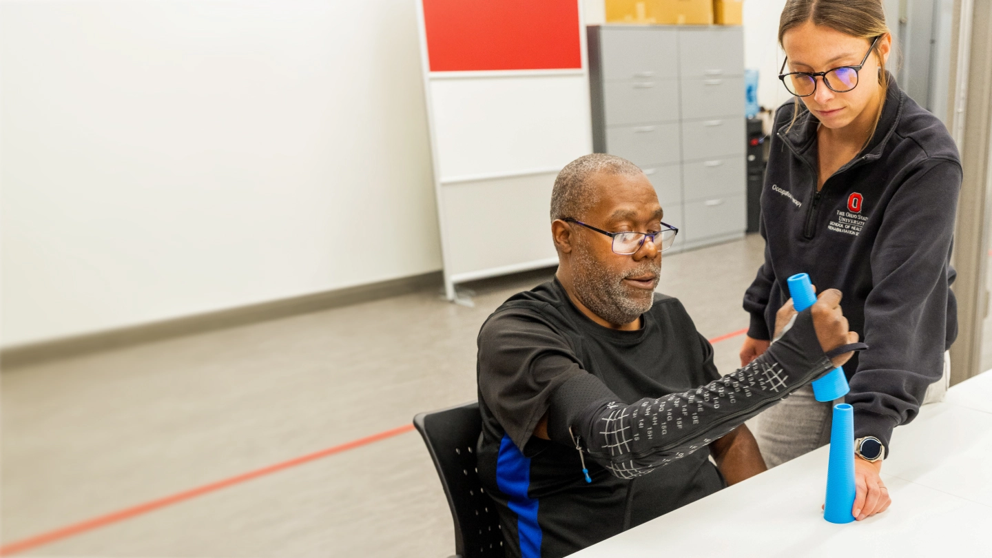 Therapist assists seated man doing hand exercises with blue cones during occupational therapy session.