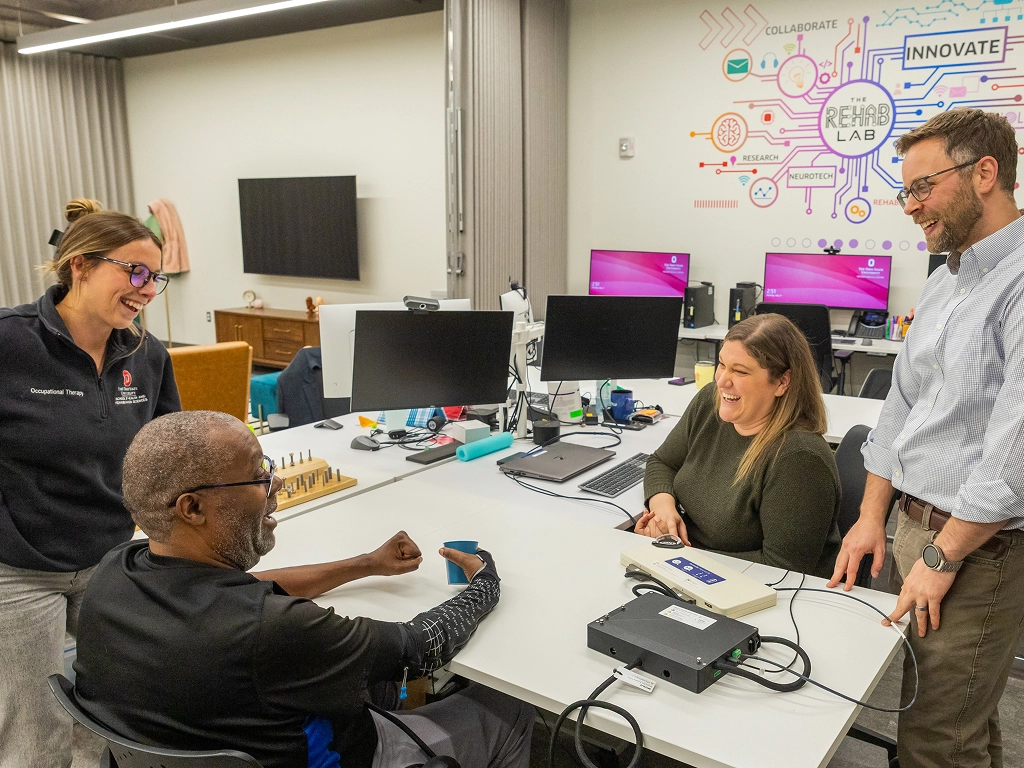 Four people smiling and talking around a table with computer equipment in a lab with innovation-themed wall art.