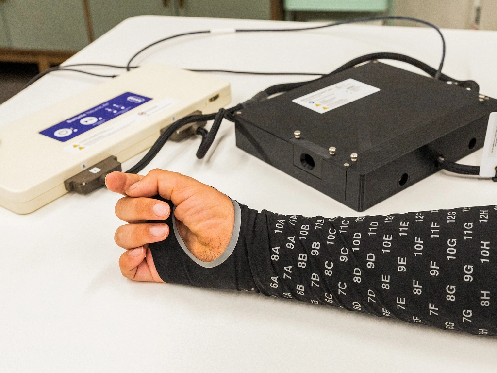 Arm with a black sleeve with white alphanumeric markings connected by wires to electronic devices on a table.