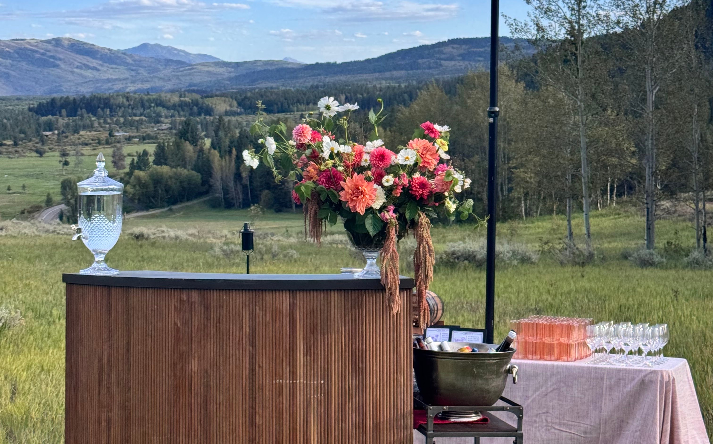 A wooden bar in the meadow with a tall arrangement of pink and coral dahlias and cosmos, aspens and the mountain ridgeline beyond.