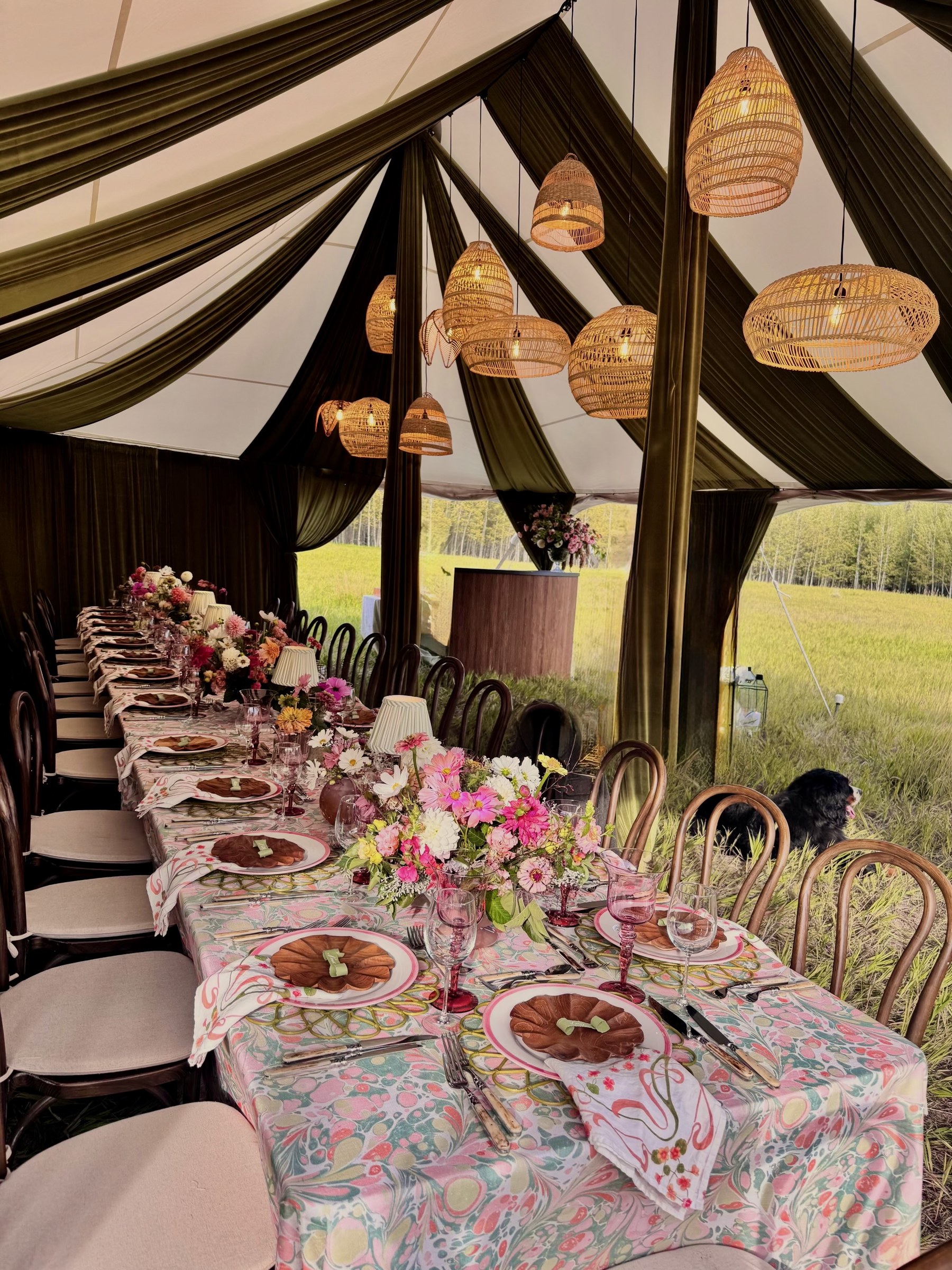 One long table under an olive-draped tent, set with marbled linens, pink-rimmed plates, wooden salad plates and wildflowers beneath woven rattan pendants, the meadow and aspens beyond.