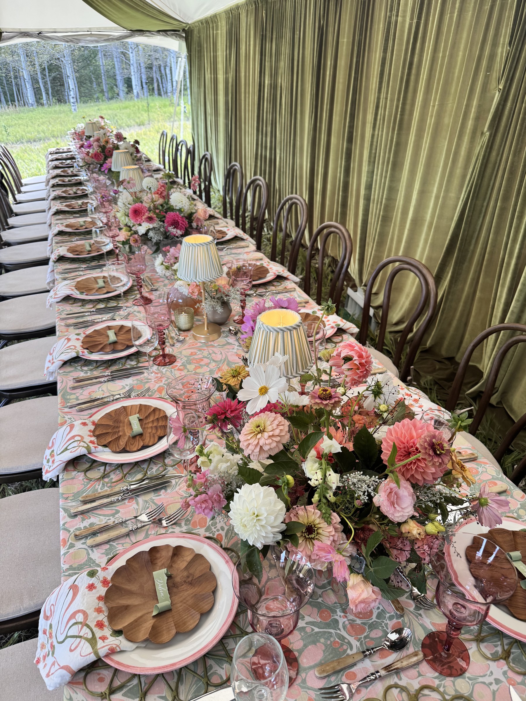 The finished long table under the tent: marbled linens, pleated lamps, pink-rimmed plates, scalloped wooden salad plates and overflowing dahlias, bentwood chairs along olive velvet drapery, the meadow grass beyond.