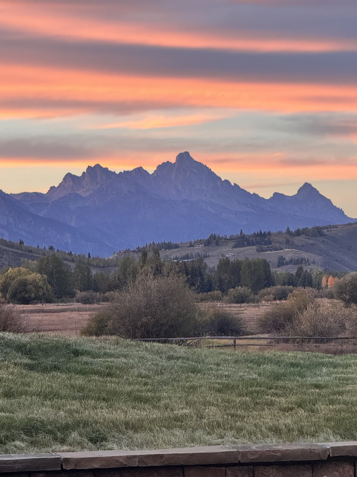 A painted sunset sky in bands of coral, pink and lavender over the Teton peaks, with cottonwoods, a rail fence and meadow grass in the foreground.