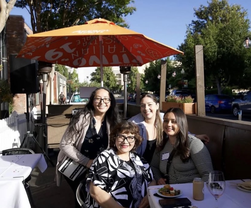 Four women smiling at an outdoor patio table under an orange umbrella on a sunny day.
