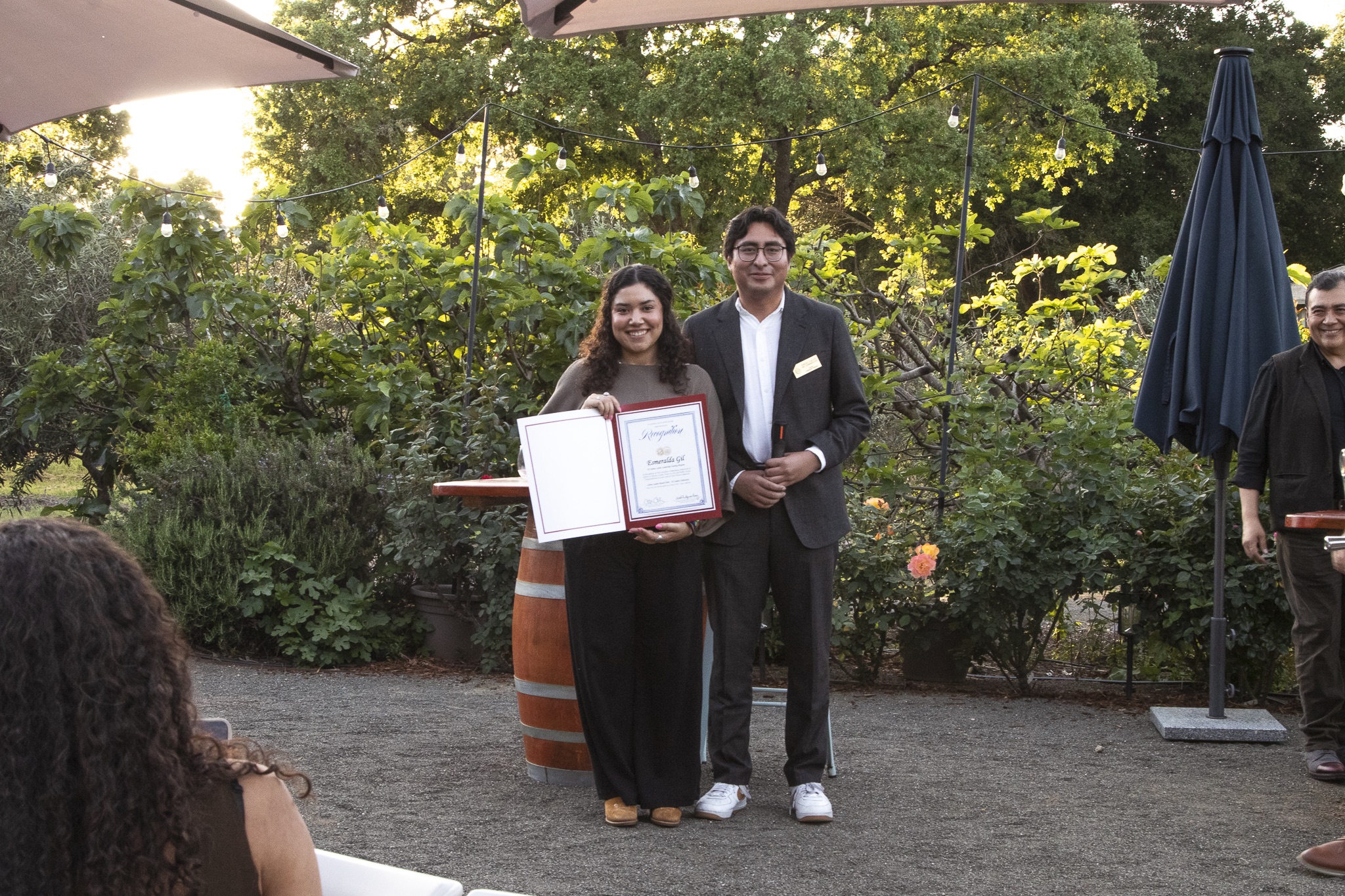 Woman holding certificate stands beside man in suit outdoors with greenery and lights in background.