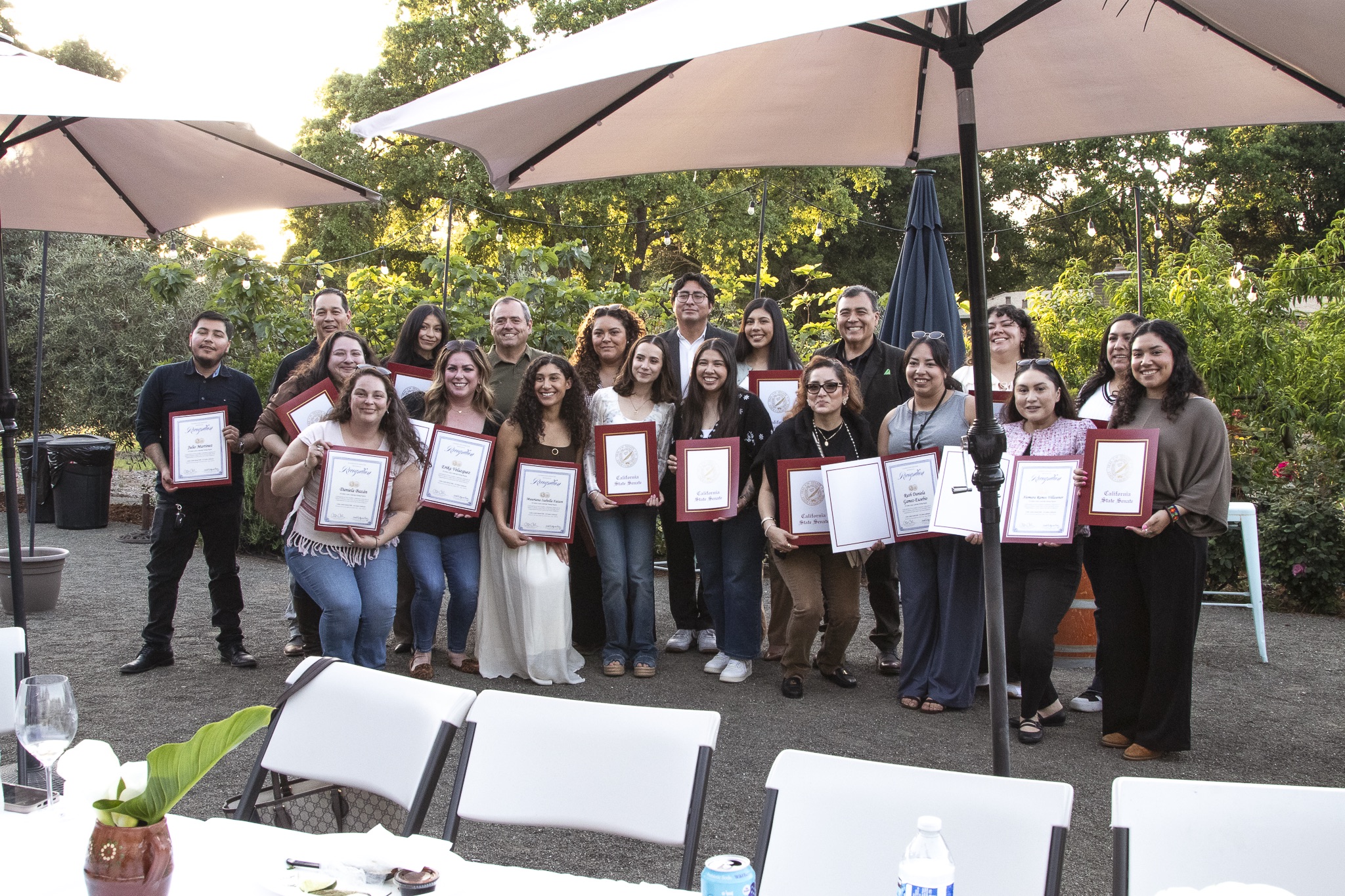 Group of people outdoors holding certificates, posing under large umbrellas with greenery in background.