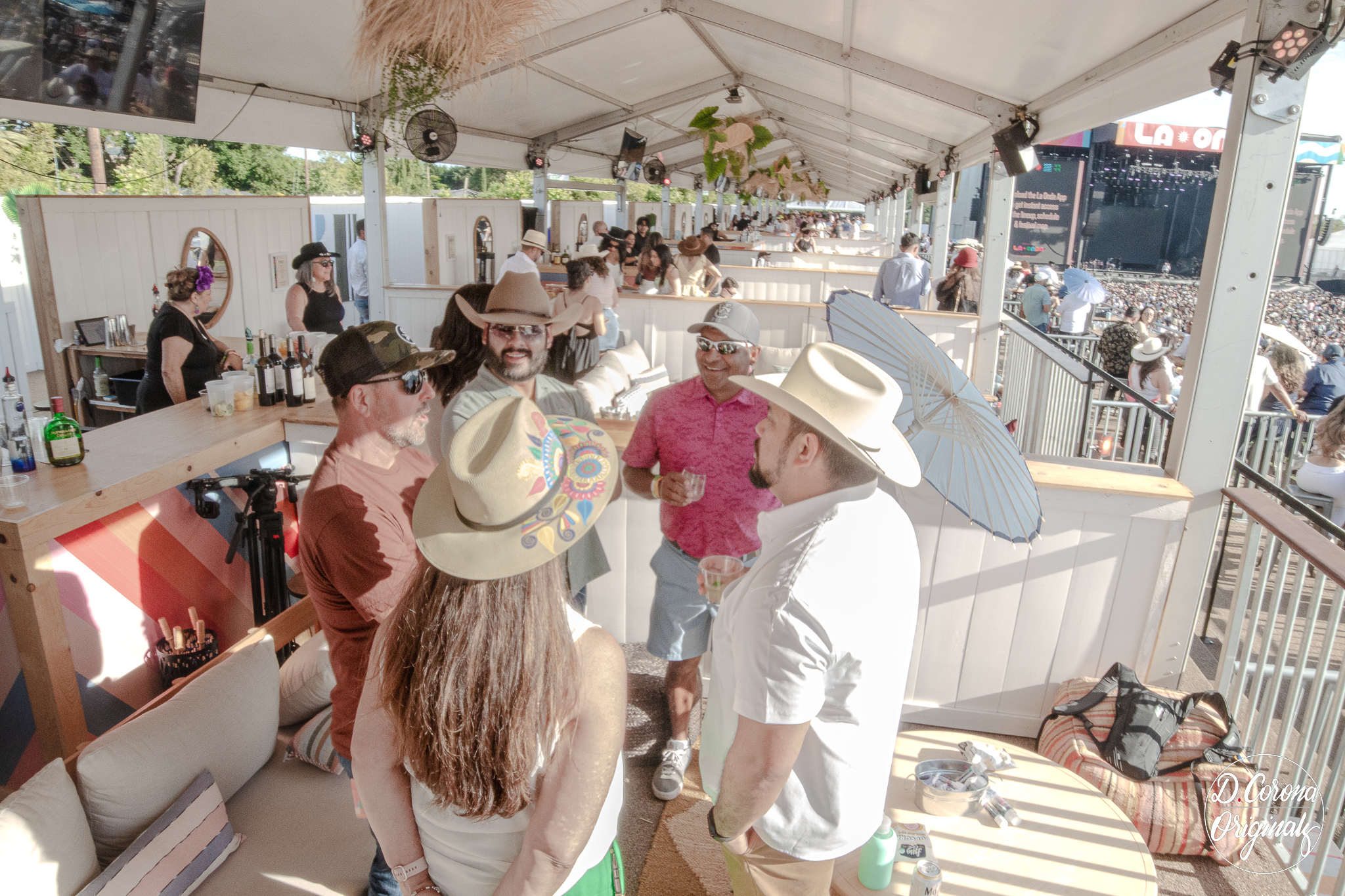 Group of five people wearing hats and sunglasses talking under a tent at an outdoor event.