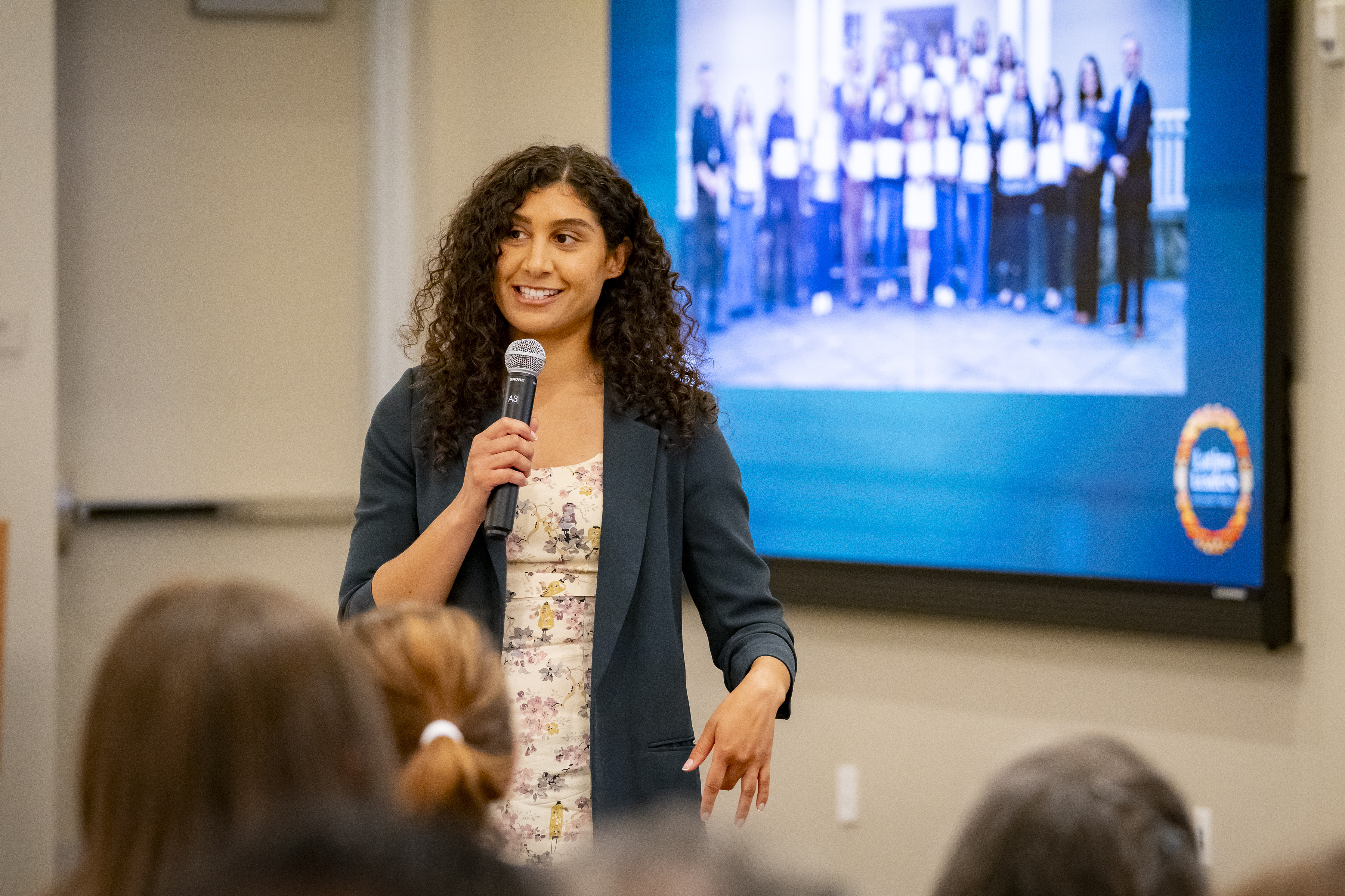 Woman with curly hair speaking into microphone in front of an audience and screen with group photo.
