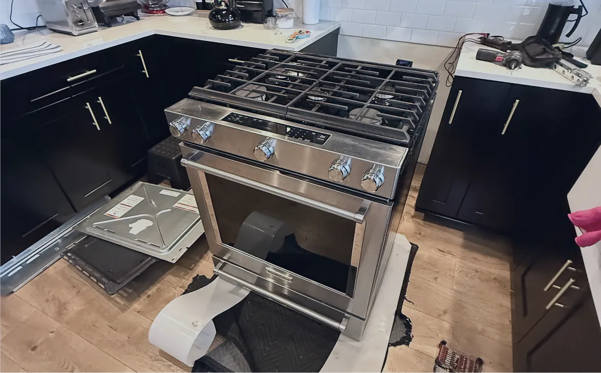 Stainless steel gas stove with oven in a kitchen surrounded by dark cabinets and countertop, with tools and parts on the floor indicating appliance repair in progress.
