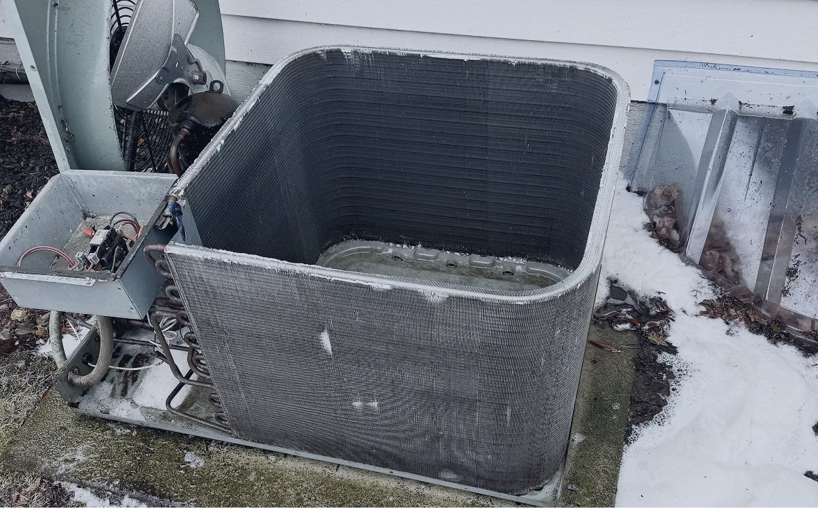 Outdoor air conditioning unit with exposed coil and electrical components, surrounded by snow and leaves.