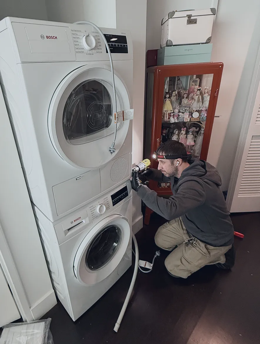 A man with a headlamp and gloves is kneeling while repairing a stacked Bosch washer and dryer unit near a red cabinet with dolls.