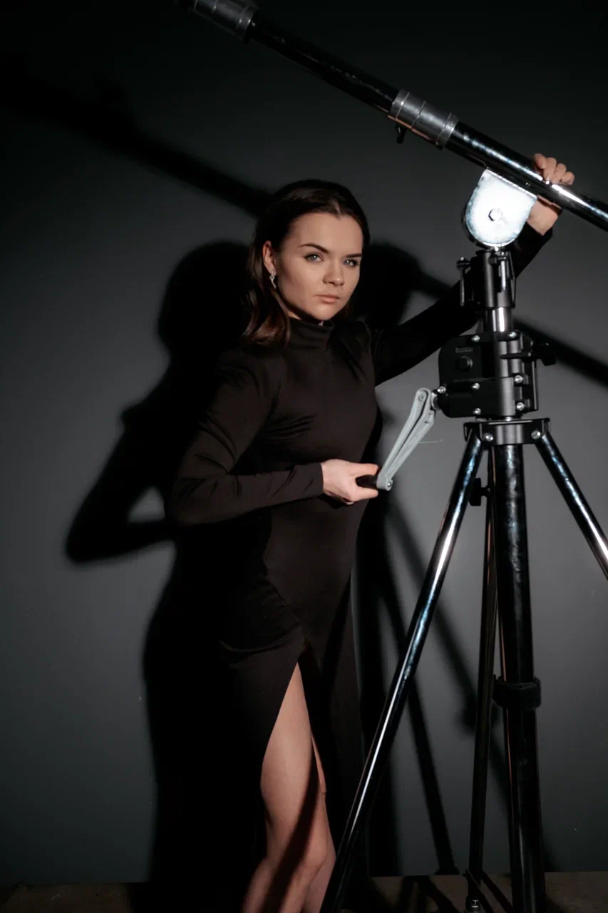 Woman in a black dress adjusting a large telescope on a tripod against a dark background.