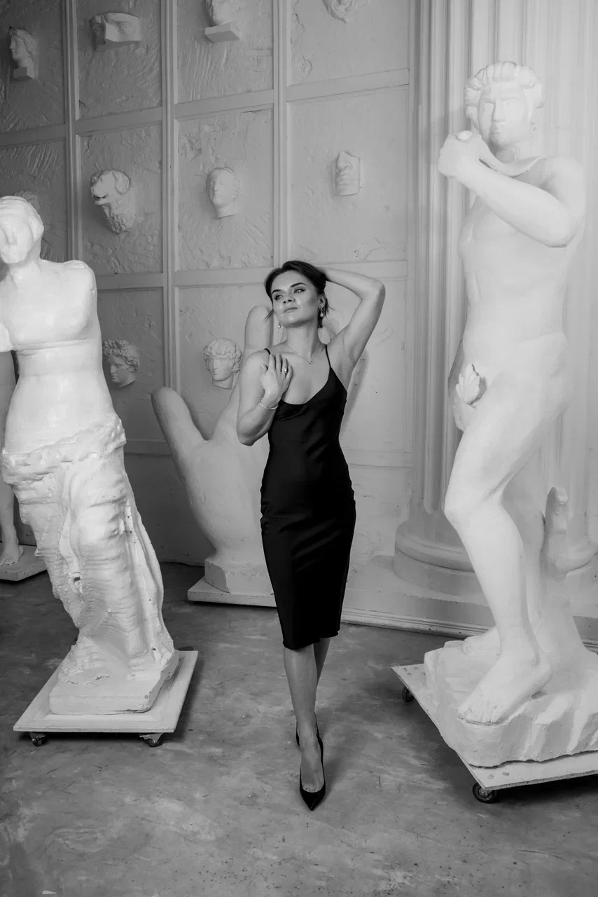 Woman in a black sleeveless dress posing between classical sculptures in an art gallery.