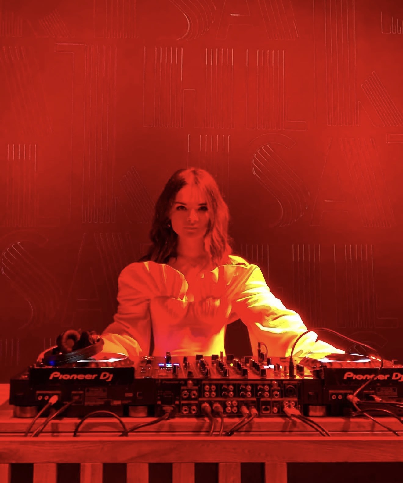 Female DJ standing behind a Pioneer DJ mixer setup under red lighting, wearing a white puff-sleeve top.