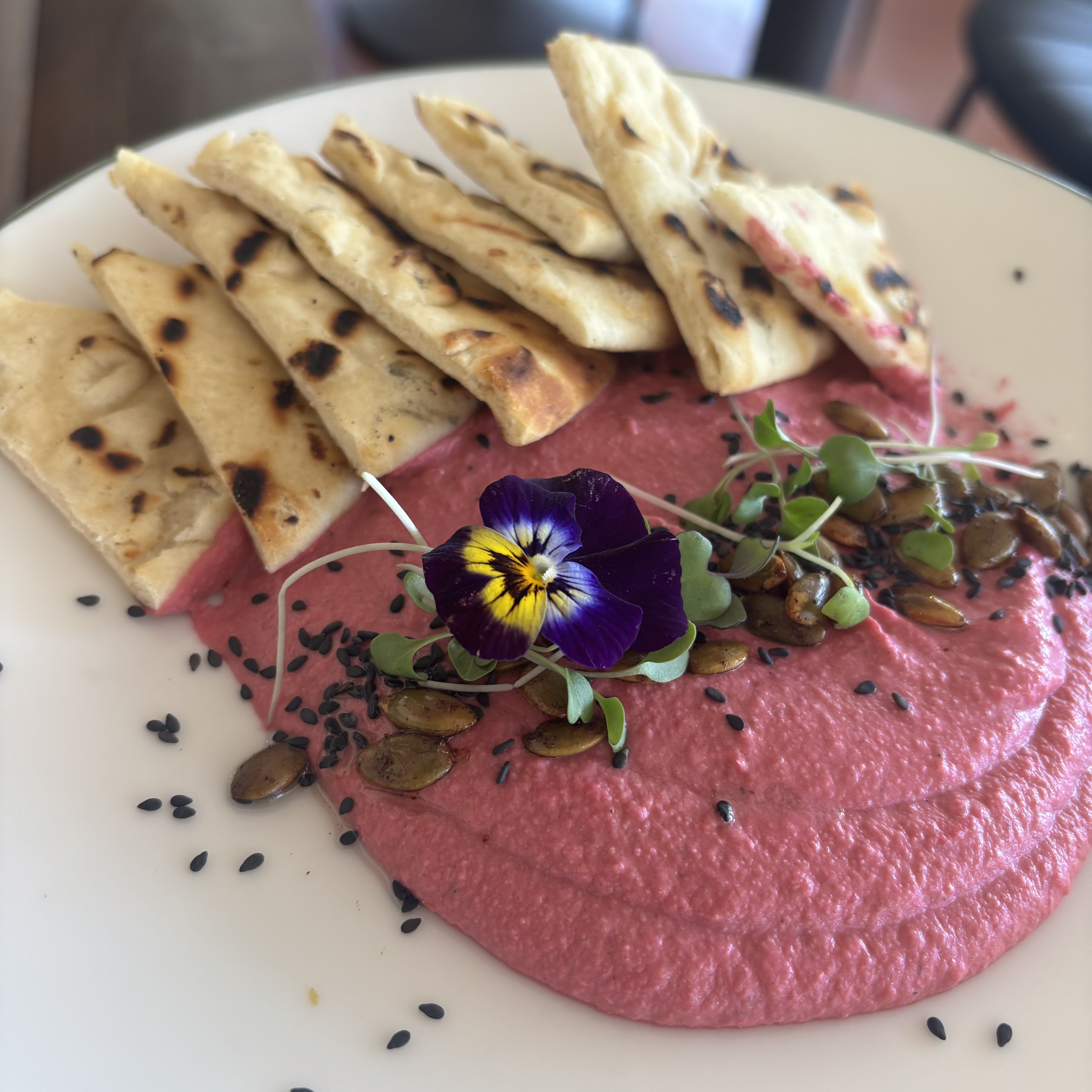 Plate of beet hummus garnished with microgreens, edible purple and yellow flower, pumpkin seeds, black sesame seeds, and served with grilled flatbread pieces.