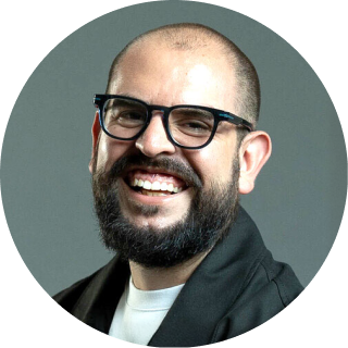 Circular profile photo of a smiling man with a full beard and dark-framed glasses, wearing a white t-shirt under a dark jacket, against a gray background.