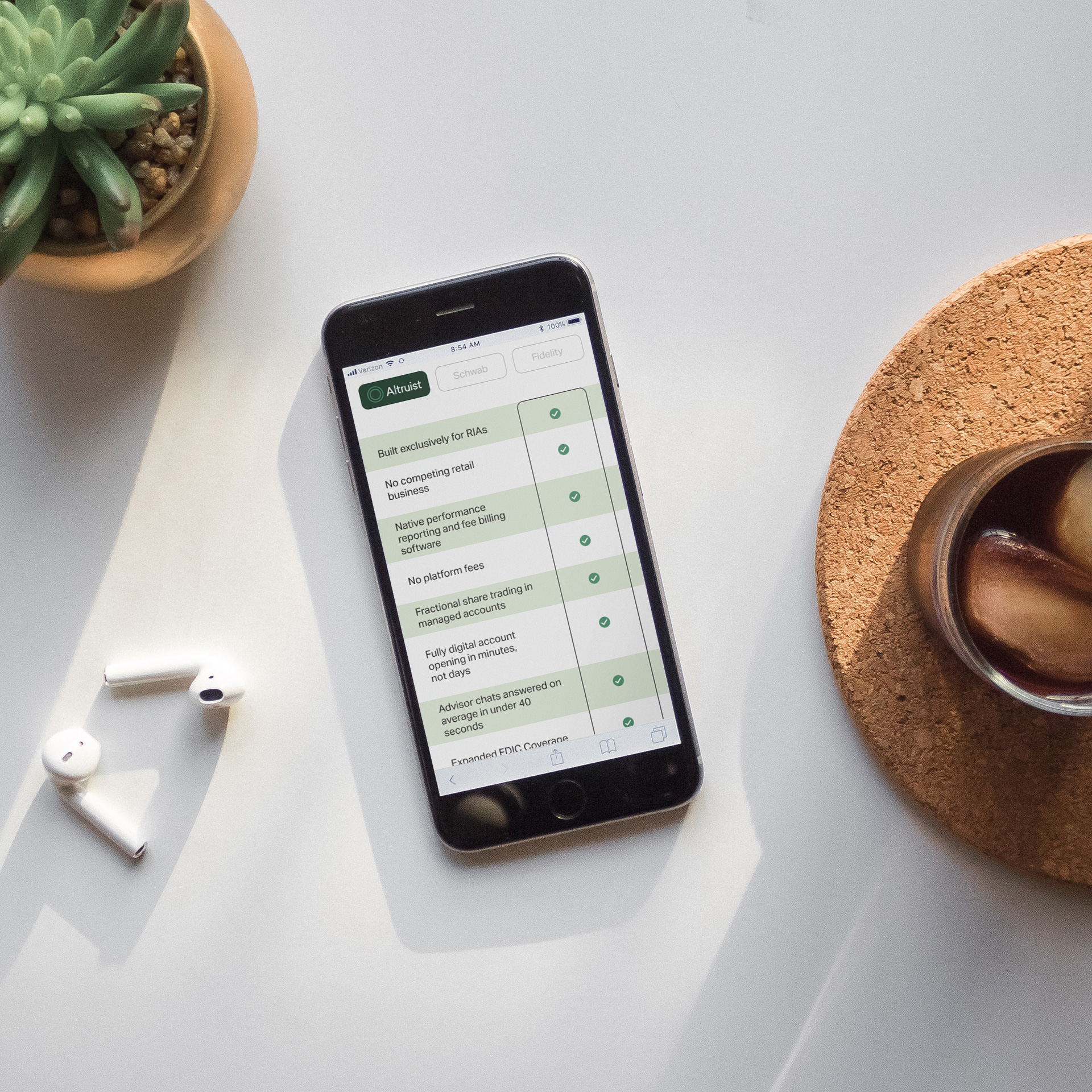Smartphone displaying a comparison chart for financial services on a white desk with earbuds, plant, and iced coffee.