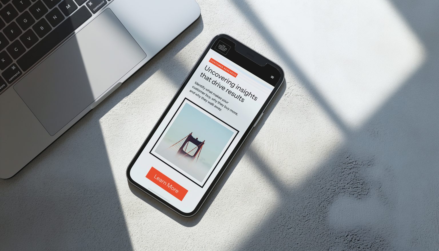 Smartphone displaying a customer research page with a bridge photo and a Learn More button beside laptop keyboard.