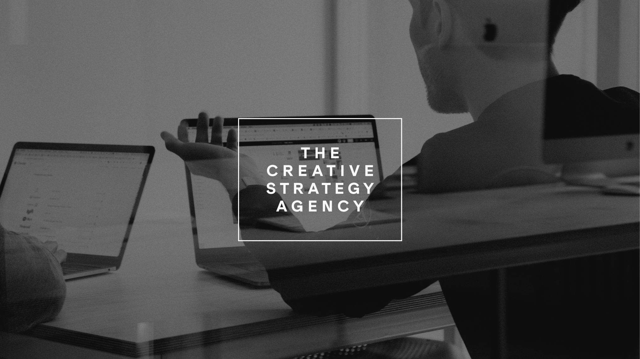 Two people working on laptops at a desk with overlay text 'The Creative Strategy Agency'.