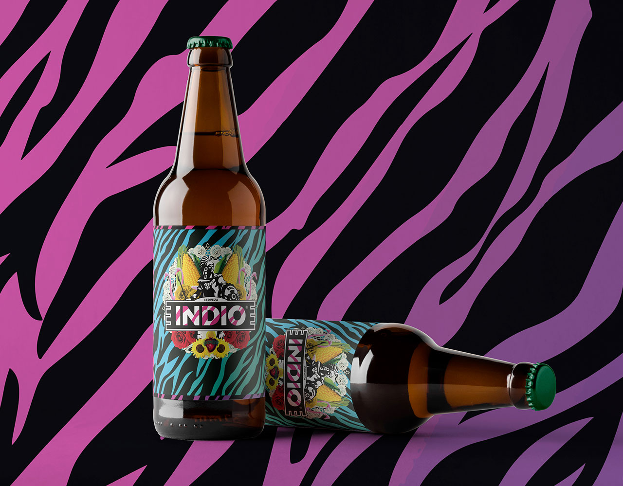 Two glass bottles of Indio beer, one upright and one lying down, showing the label redesign with a corn-and-skeleton motif, roses, and sunflowers, set against a pink and black zebra-print background.