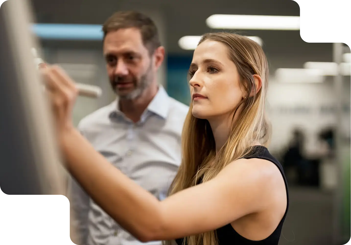 Focused woman writing on a transparent board with a marker while a man watches attentively behind her in an office setting.