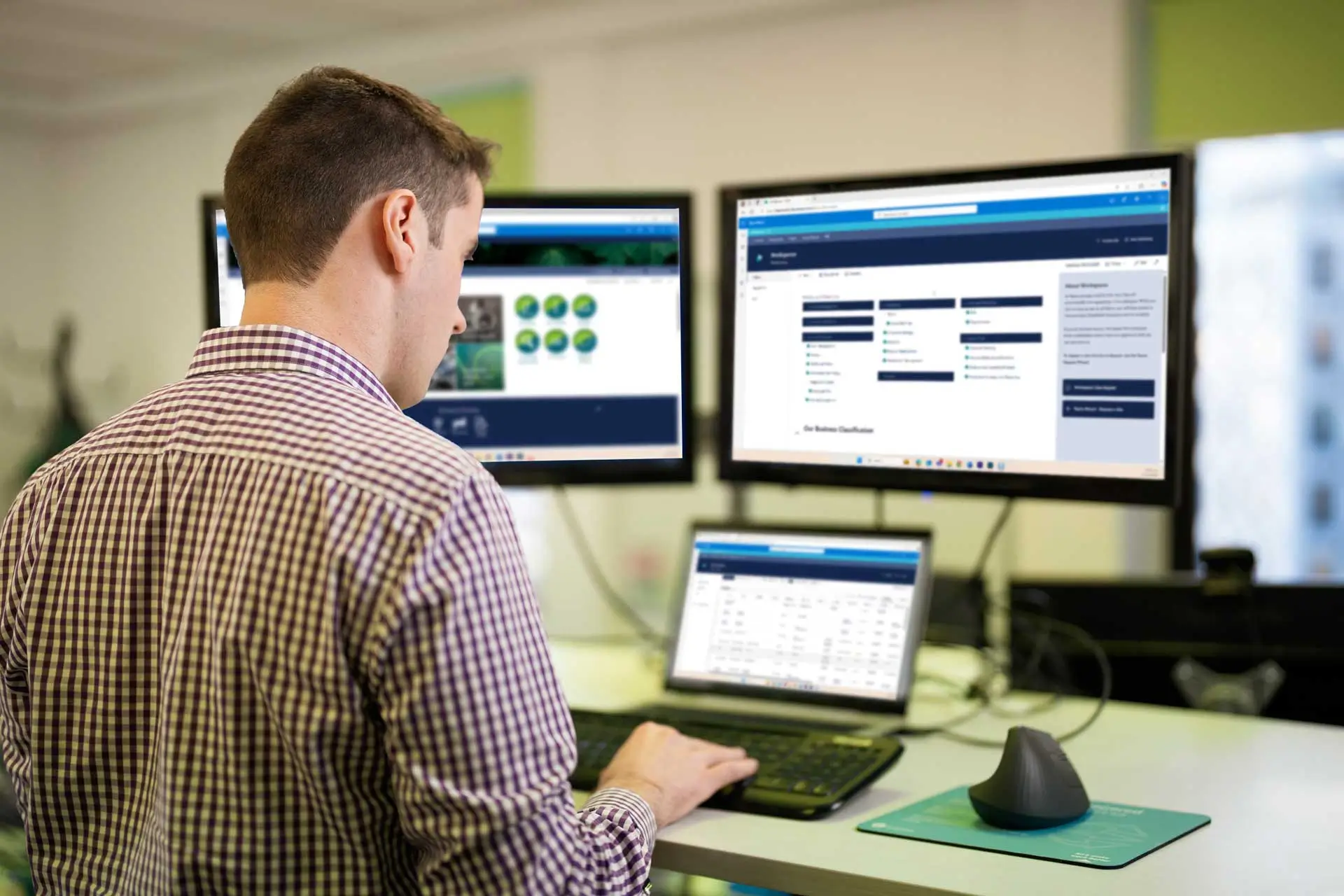 Man in checkered shirt working at desk with laptop and two monitors displaying data and charts.