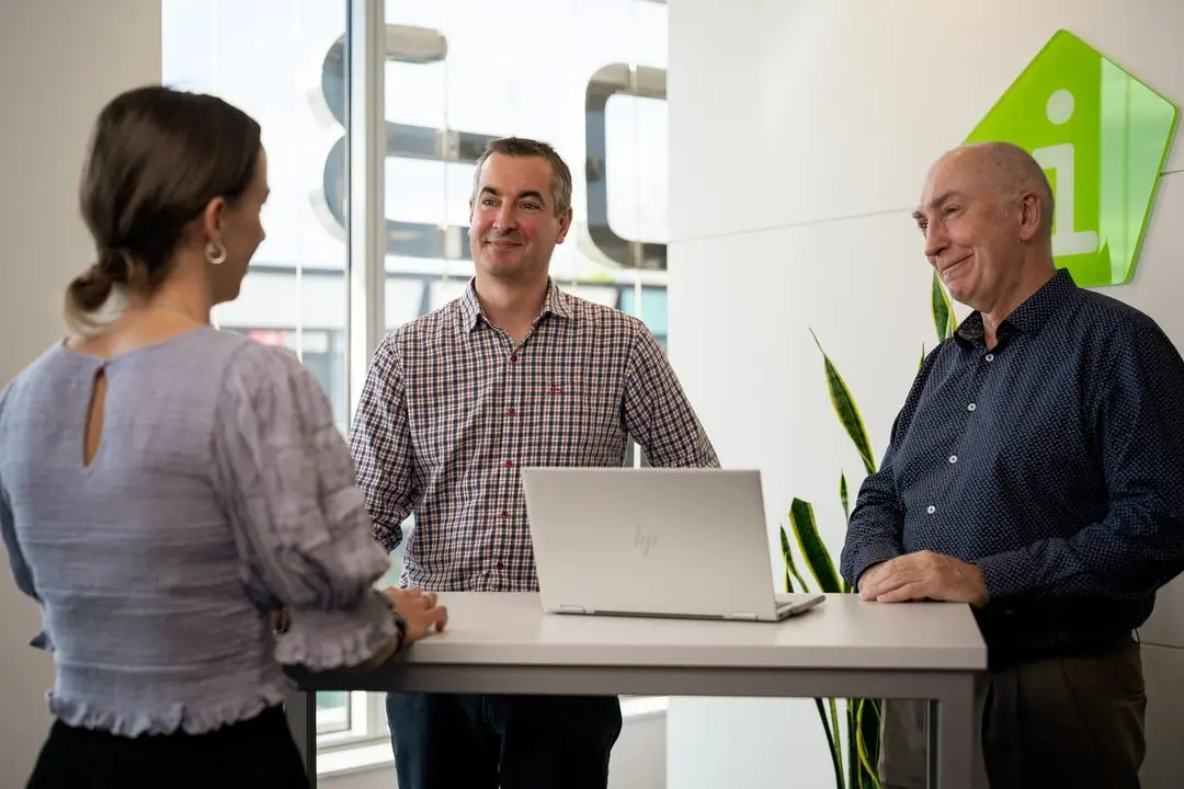 Three people talking at a standing desk with a laptop in a modern office setting.