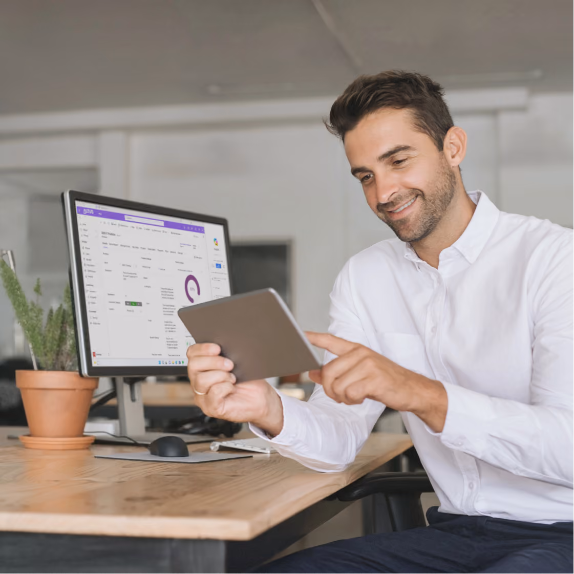 Smiling man in white shirt using a tablet at a desk with a computer and potted plant.