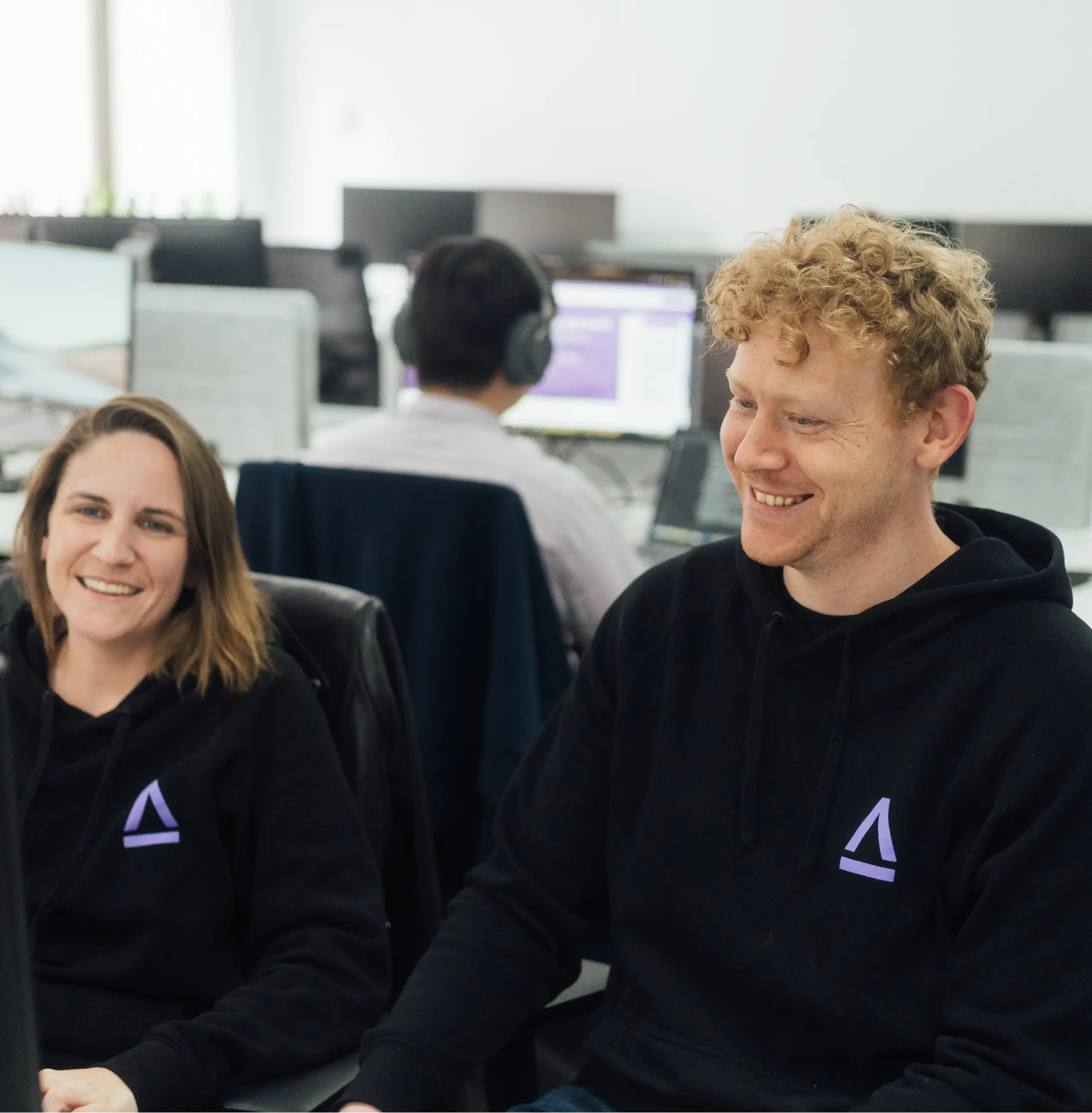 Two smiling coworkers wearing matching black hoodies sitting together in a modern office space.