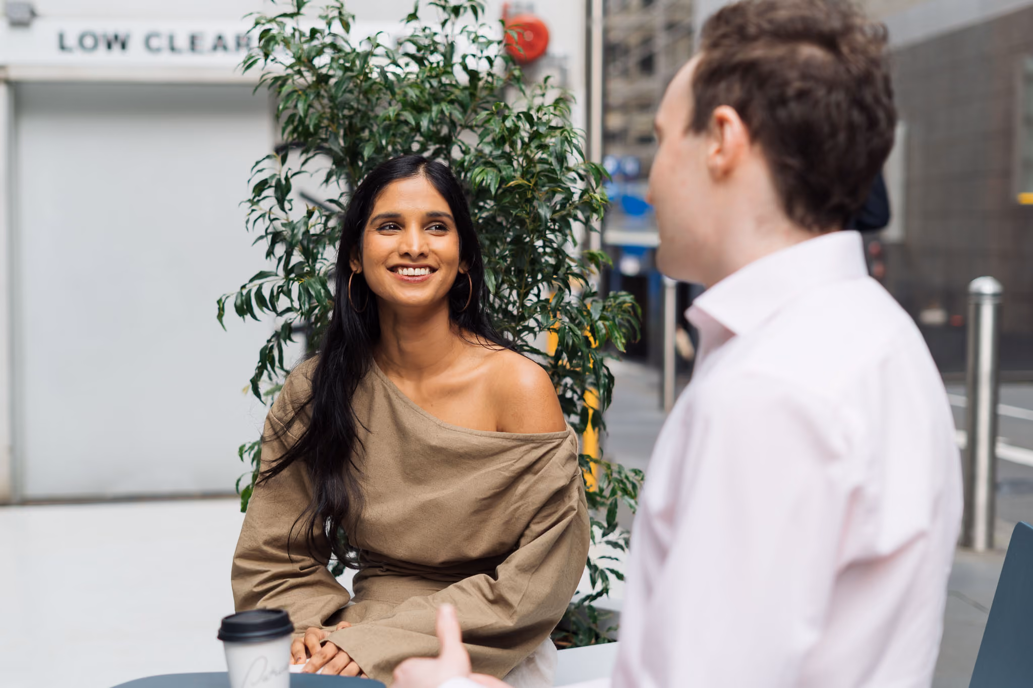 Smiling woman in tan off-shoulder top talks with man in white shirt outdoors near coffee cup.