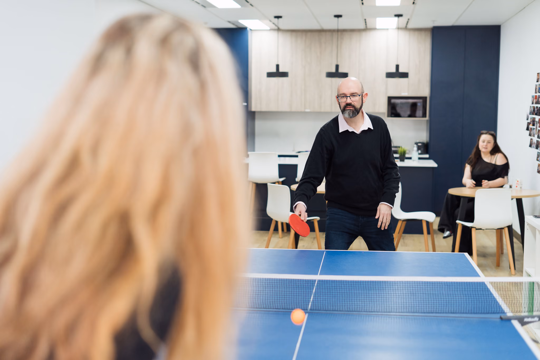 Man with glasses playing table tennis in an office while a woman watches from a nearby table.