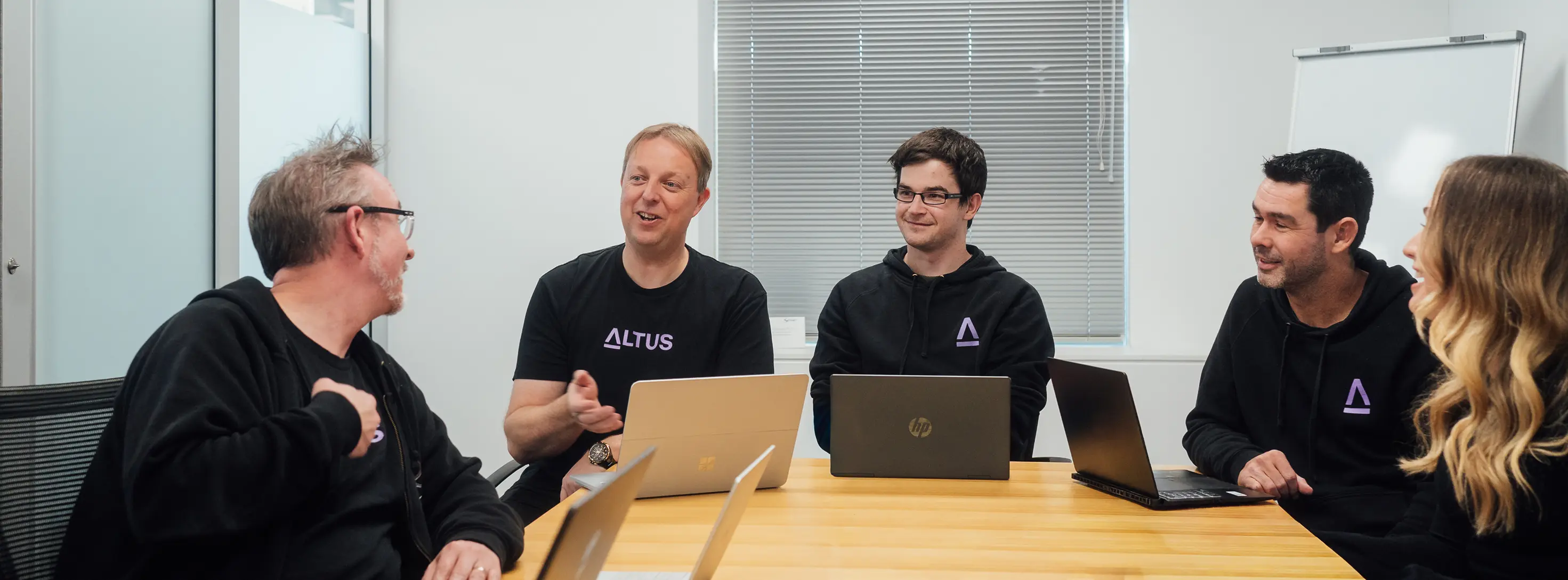 Five colleagues wearing black Altus shirts having a meeting with laptops at a wooden table.