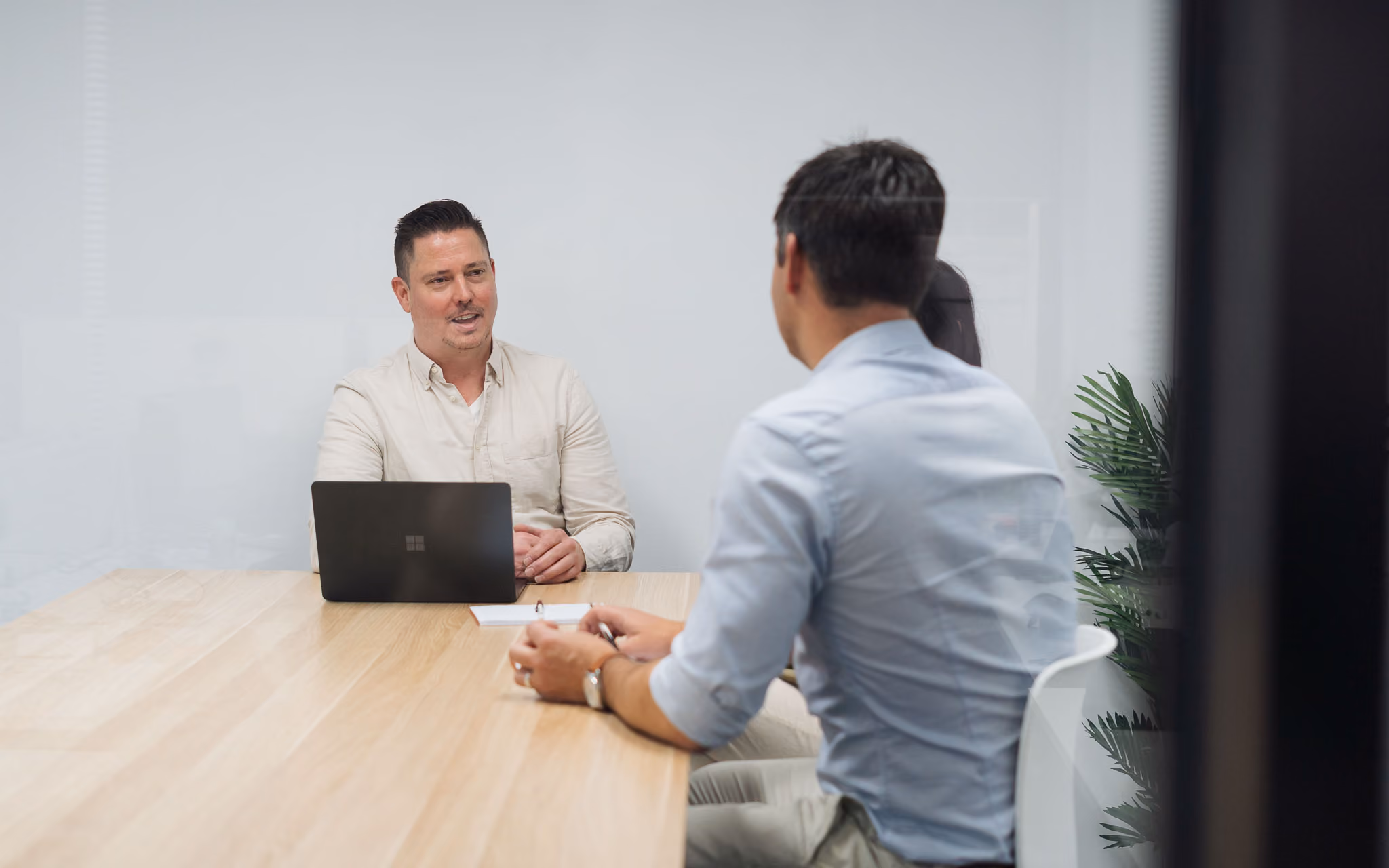 Two men having a meeting across a wooden table, one with a laptop and the other holding a pen.
