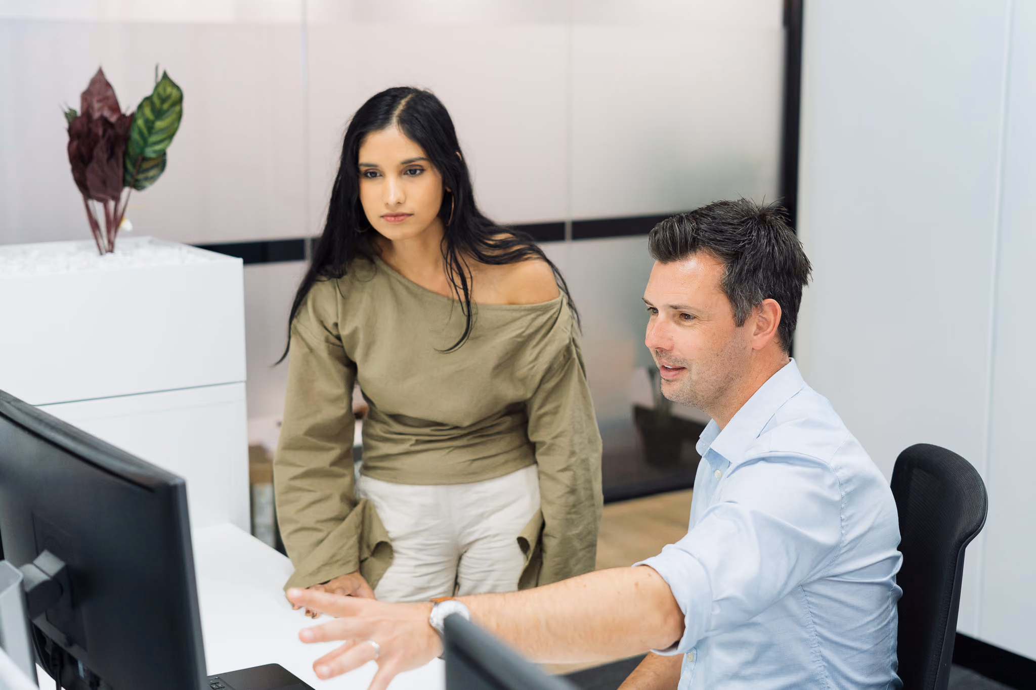 Man seated pointing at a computer screen with a standing woman watching attentively in an office.
