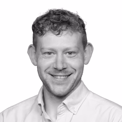 Smiling man with curly hair wearing a buttoned light-colored shirt against a white background.